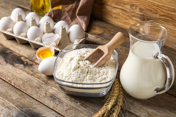 Bowl with flour, eggs and jug with milk on wooden table
