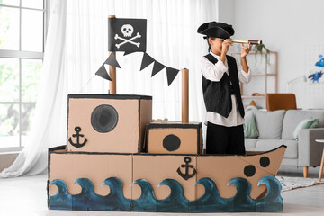 Little boy dressed as pirate playing with spyglass and cardboard ship at home - obrazy, fototapety, plakaty