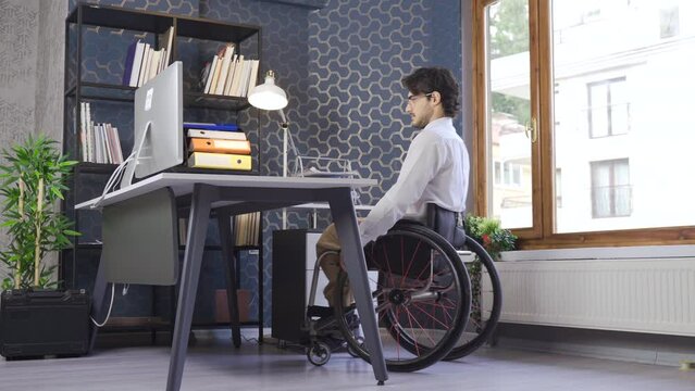 Motivated Disabled Person Self Employed Working in Office.
Disabled businessman Sitting in Wheelchair comes to his desk and Working on Computer Typing.
