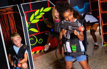 Fototapeta na wymiar Smiling happy cheerful little girl aiming laser gun at other players during lasertag game in dark room.