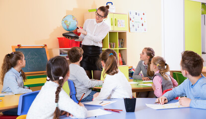 Cheerful positive female teacher giving geography lesson in classroom, showing pupils globe