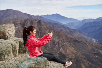 Fototapeta premium young woman sideways taking a picture with her smartphone, wearing a red jacket, sitting on a rock in the middle of the Andes mountain range in Chile