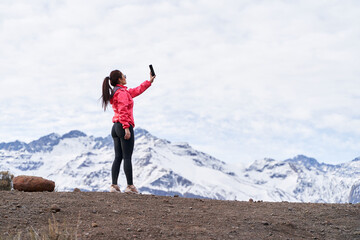 young woman standing in red jacket, taking a picture with her phone in the middle of the Andes Mountains of Chile