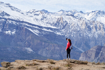 Naklejka na ściany i meble young red haired woman with red jacket and backpack, taking pictures with her phone in the middle of the Andes mountain range in Chile