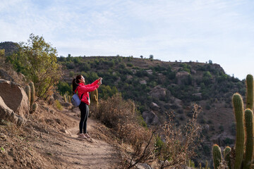 Fototapeta premium young red haired woman with red jacket and backpack, taking pictures with her phone in the middle of the Andes mountain range in Chile