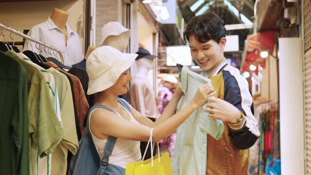 4K Young Asian couple enjoy and fun outdoor lifestyle shopping together at street market on summer holiday vacation. Happy man and woman choosing and buying fashion clothes together at clothing shop.