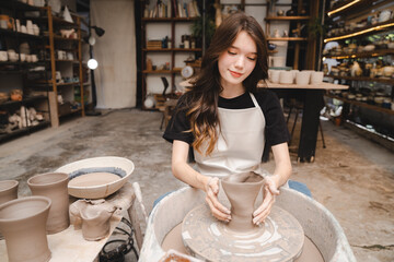 Beautiful young woman holding pottery instrument for scraping, smoothing, shaping and sculpting. Lady siting on bench with pottery wheel and making clay pot - obrazy, fototapety, plakaty