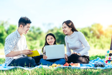 Happy asian family in the garden, Father, Mother and doughter. They are having fun playing song with ukulele, reading book, using laptop in holiday.