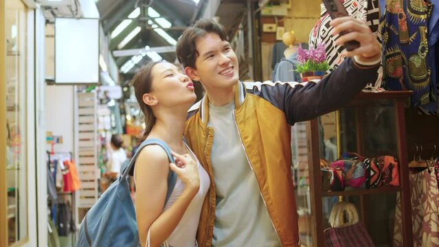 4K Asian couple enjoy and fun outdoor lifestyle shopping at street market on summer holiday vacation. Man and woman couple using mobile phone taking selfie together while shopping at weekend market.