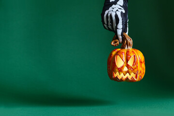 Skeleton gloved hand holds ceramic pumpkin jack-o'-lantern on green background