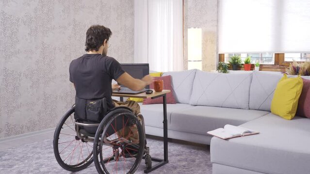 Disabled young man sits in wheelchair and works with laptop.
Disabled teenager in wheelchair working with laptop at home.
