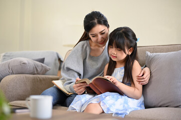 Lovely and relaxed Asian mom reading a book or telling fairy tale story to her cute daughter