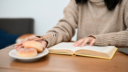 A smart Asian female college student eating doughnut while reading a book. cropped image
