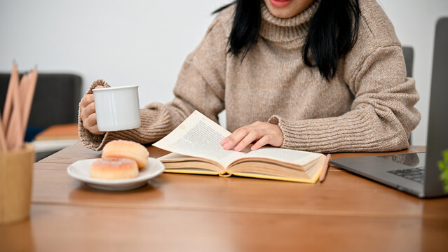 Asian Female In Comfy Sweater Relaxes While Reading A Book And Sipping Hot Cocoa.