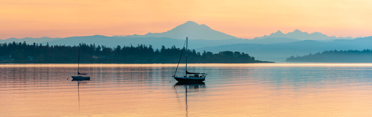 Colorful Sunrise Over Mt. Baker With a Sailboat in the Foreground.  Beautiful calm morning in the San Juan Islands as the majestic Mt. Baker looms in the background. - obrazy, fototapety, plakaty
