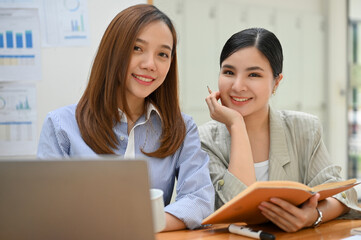 Two Asian businesswomen sits at office desk, smiling and looking at the camera.