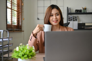 Beautiful millennial Asian businesswoman looking at laptop screen while sipping morning coffee.
