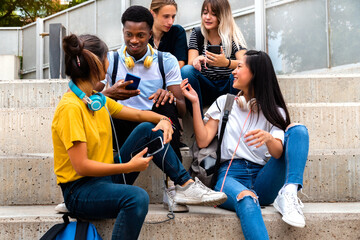 Happy multiracial teenage high school students sit on steps talking and using mobile phones outside.
