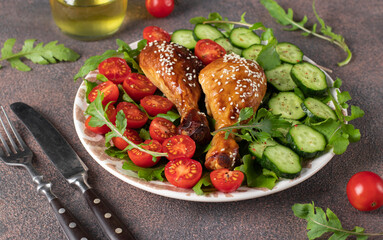 Baked chicken drumsticks with cherry tomato, cucumber and arugula salad on brown background