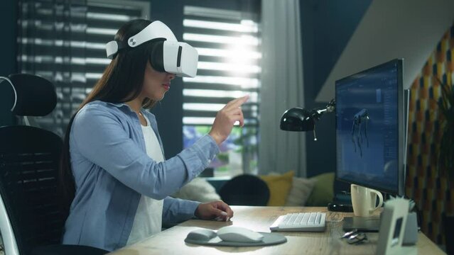 Asian woman sitting at the table at home office and using VR headset to work on 3D modeling project on personal computer. Virtual reality. Metaverse