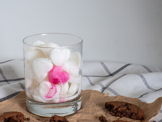 White Ice cubes made from milk in heart shape with one red heart in glass. Space for text. Cocoa cookies with chocolate on paper on a white background. Valentines day breakfast eating, love concept.