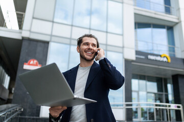 Fototapeta premium Satisfied Man speaks on a mobile phone with a laptop in his hands on the background of a business center, mobile operator concept