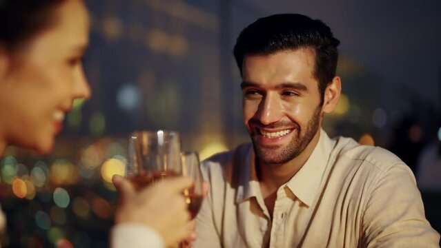 4K Caucasian couple celebrating holiday event at luxury skyscraper rooftop bar at night. People enjoy city nightlife having dinner and drinking wine at outdoor rooftop restaurant on summer vacation.