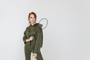 attractive young red-haired woman in a green tracksuit holding a tennis racket on a white background copy space