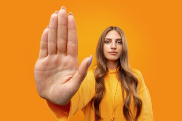 Young woman standing with outstretched arm showing stop sign against yellow background - obrazy, fototapety, plakaty