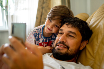 A smiling young man with a beard in casual clothes lying on the sofa in the living room and using a mobile phone. The son is standing next to him and looks at his father on the phone