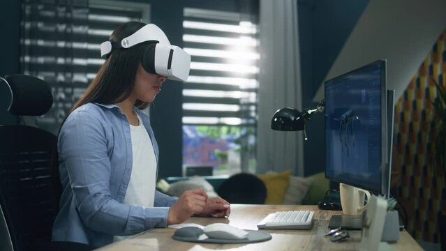 Asian female 3D designer in VR goggles working on pc in 3D modeling program remotely from home office and creating 3D prototype of modern robot. Virtual reality. Metaverse