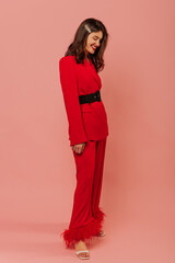 Full-length, modest young caucasian girl smiles with her eyes closed standing sideways on pink background. Brunette with wavy hair is wearing red pantsuit and white sandals.