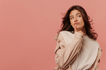 Young caucasian girl looks pensively sideways with her finger on chin against pink background with place for text. Brunette girl with wavy hair wears light-colored casual clothes.