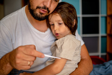 A sick child sits on the father's lap. Dad takes his son's temperature