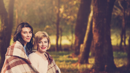 Two smiling women wrapped in blanket posing in autumn forest with baby carriage