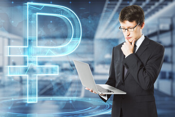 Attractive young european businessman with laptop and glowing ruble and map hologram on blurry office interior background. Money, currency, trade, online banking and app concept. Double exposure.