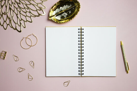 A white notebook with golden leave, earrings, and pen over the pink background. 