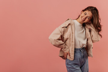 Fashionable young caucasian girl with smile on her face poses on pink background with place for text. Brunette girl wears light-colored casual clothes. Lifestyle, different emotions, leisure concept. 