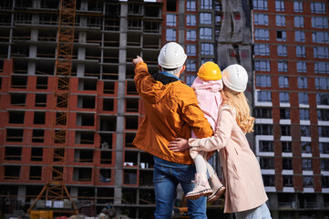 Obraz na płótnie Canvas Back view of man pointing at apartment building under constructing while standing next to wife and daughter on the street. Happy family looking at new home at construction site.