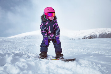 Fototapeta na wymiar Little girl snowboarder on empty track at ski resort in sunny winter day. Portrait of kid in sportswear with equipment ride on slope