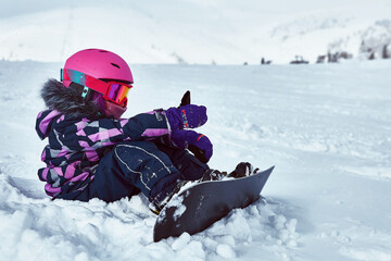 Little girl snowboarder sitting on piste at ski resort in sunny winter day. Portrait of kid in sportswear with equipment resting on ski slope