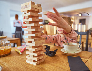 Kid girl playing stacking wood blocks (Jenga) funny and joyful on game in cosiness coffee house. Hand movement control building computational skills children's play concept.