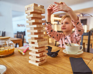 Kid girl playing stacking wood blocks (Jenga) funny and joyful on game in cosiness coffee house. Hand movement control building computational skills children's play concept.