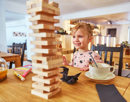 Kid girl playing stacking wood blocks (Jenga) funny and joyful on game in cosiness coffee house. Hand movement control building computational skills children's play concept.