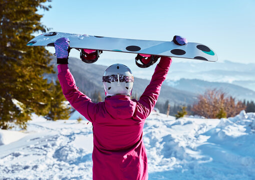 Back View Of Woman With Equipment Helmet And Goggles Outwear Holding Snowboard Over Head And Looking Mountain's Landscape In Sunlight. Mountain's Top In Sunny Winter Day. Free Space For You Text