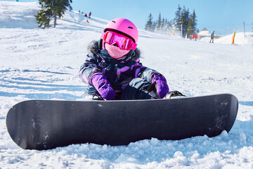 Little girl snowboarder sitting on piste at ski resort in sunny winter day. Portrait of kid in...