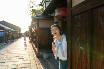 backlight shot of amazed asian Japanese female tourist popping head from behind entrance of teahouse and looking into historic street at Ninenzaka and Sannenzaka in Kyoto japan at twilight