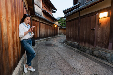 Fototapeta na wymiar full length of relaxed asian Japanese girl visitor leaning against wooden wall and looking at her cellphone in illuminated Ishibeikoji alley in gion Kyoto japan at sunset