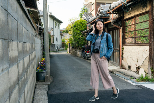 full length of stylish asian chinese girl posing with hand on her head in an alleyway at residential area in uji Kyoto japan. she stands near an old Japanese house with paints peeling off wall