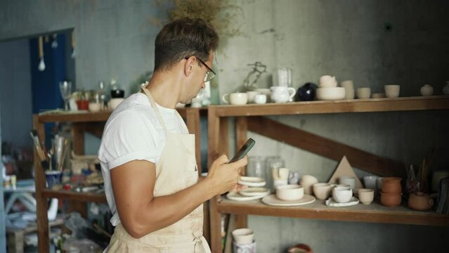 Young happy potter photographing his clay pottery using his phone in his studio. Concept of education, business, technology and creative handmade